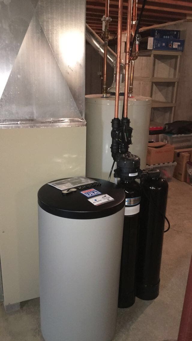 A water softener is sitting next to a water heater in a basement.