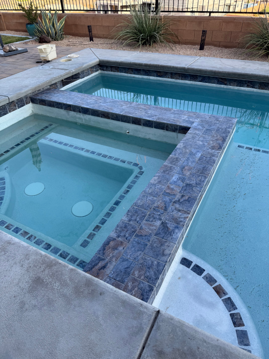 An outdoor swimming pool with an attached square hot tub, featuring blue stone tile trim and concrete decking.