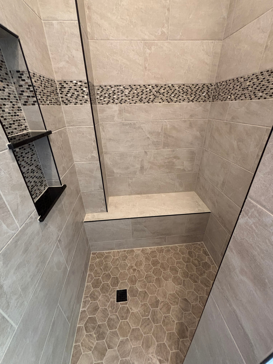 A walk-in shower with beige tiles, a built-in bench, a black-tiled shower niche, and a hexagon-tiled floor.