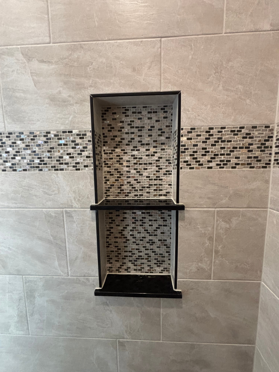 A rectangular tiled shower niche with black stone shelves and a decorative mosaic tile accent strip on a light wall.
