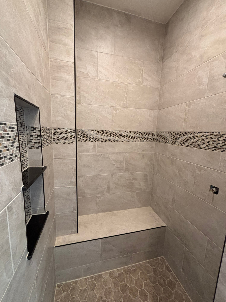 A tiled shower stall featuring a stone-look bench, a mosaic accent strip, and a recessed black-shelved wall niche.