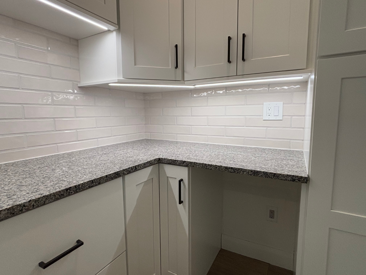 A corner kitchen counter with light granite, white cabinets, white subway tile backsplash, and under-cabinet lighting.