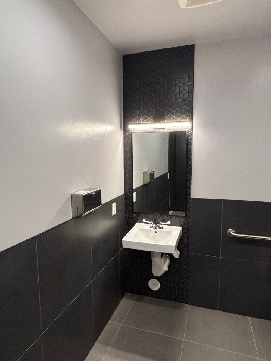 A modern, accessible public restroom with gray walls, a black-tiled accent wall, a white wall-mounted sink, and a grab bar.