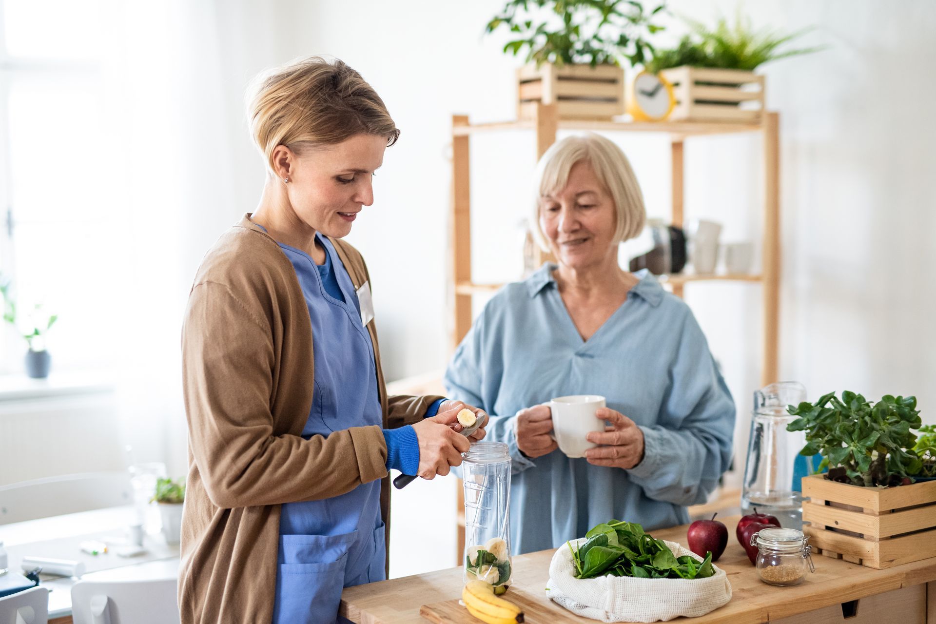 Woman assisting another woman, preparing smoothie in a kitchen.