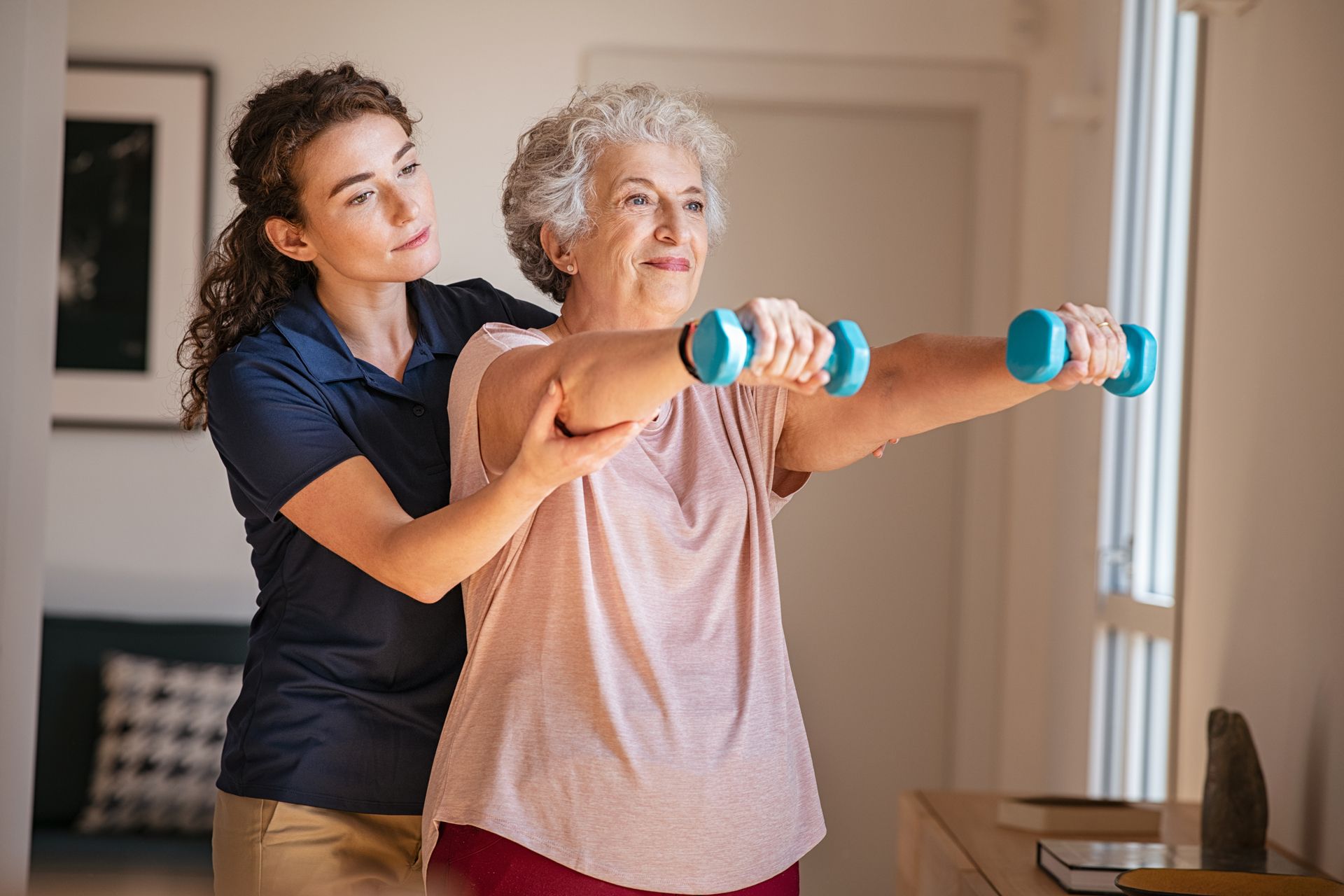 Woman assisting senior lifting weights, sunny room.