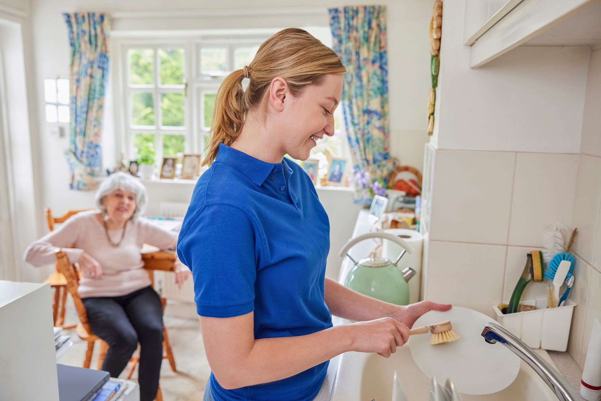 Woman washing dishes in kitchen, elderly woman seated at table nearby.