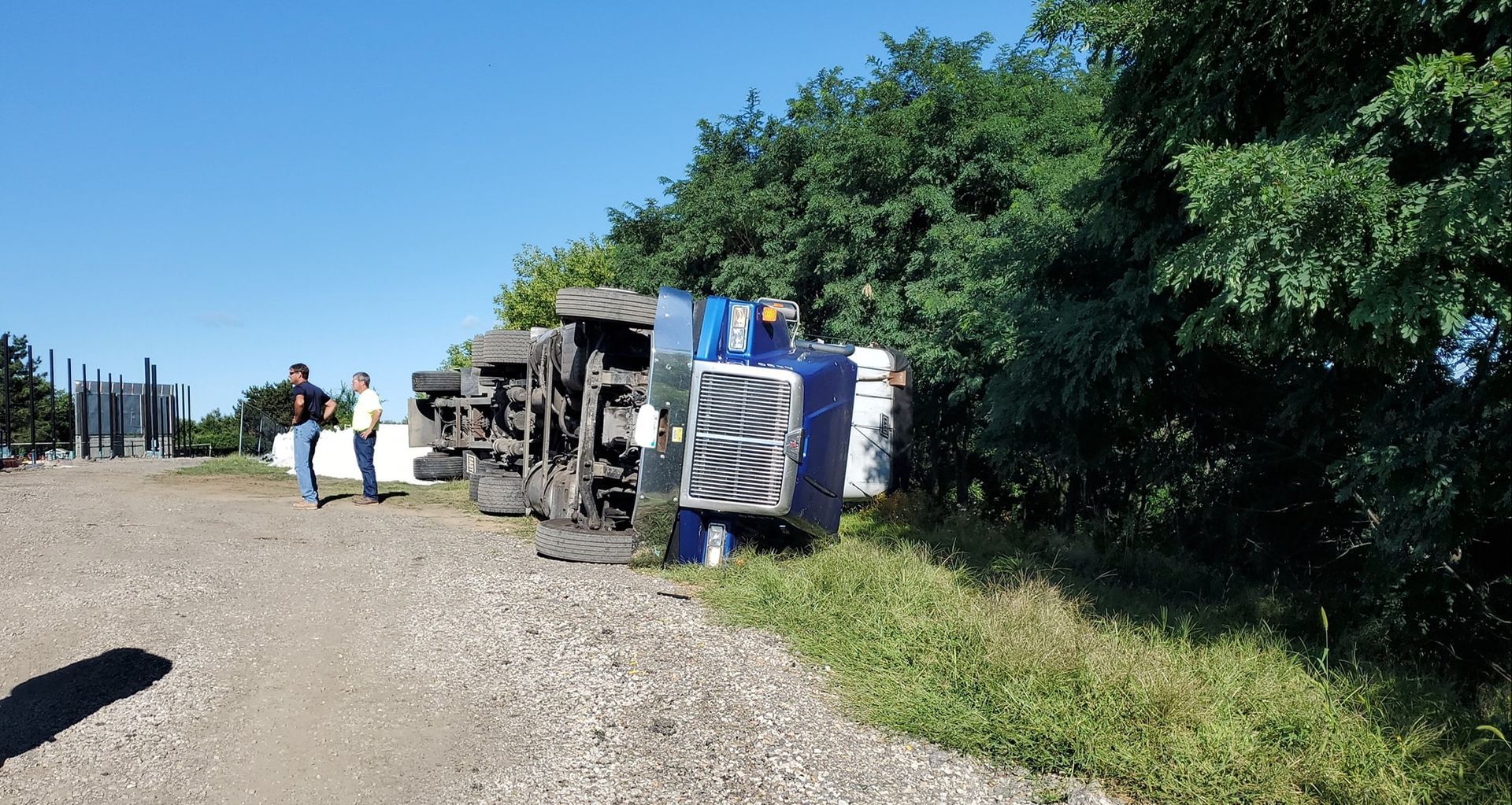 Overturned blue semi-truck on dirt road next to green foliage, two people standing nearby.