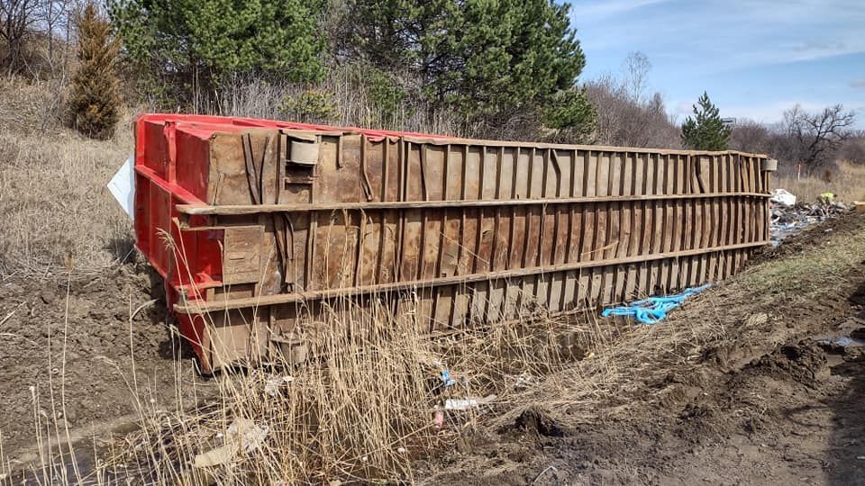 A red overturned semi-trailer in a ditch along a roadside with a wooded backdrop.