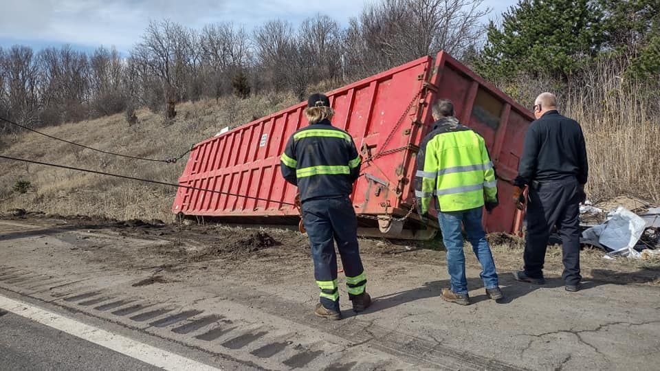 Three people assess a tipped red dumpster on the side of a road; a wooded hillside is in the background.