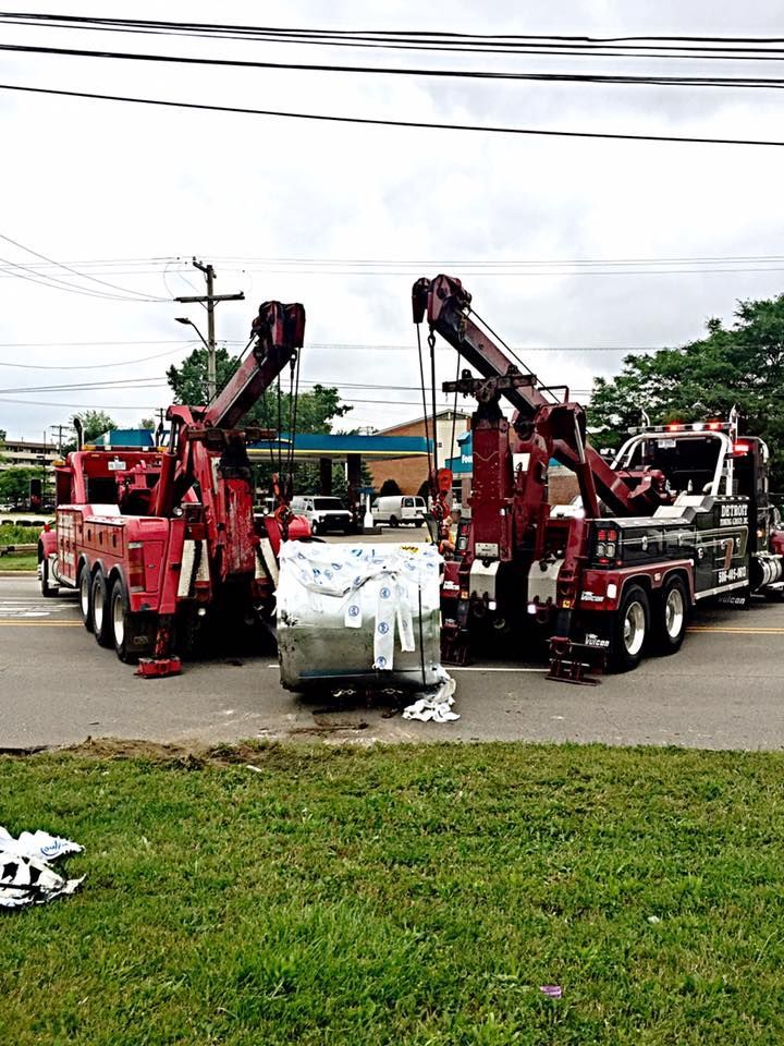 Two red tow trucks lifting a large, wrapped object in the street; grassy area in front, gas station in the background.