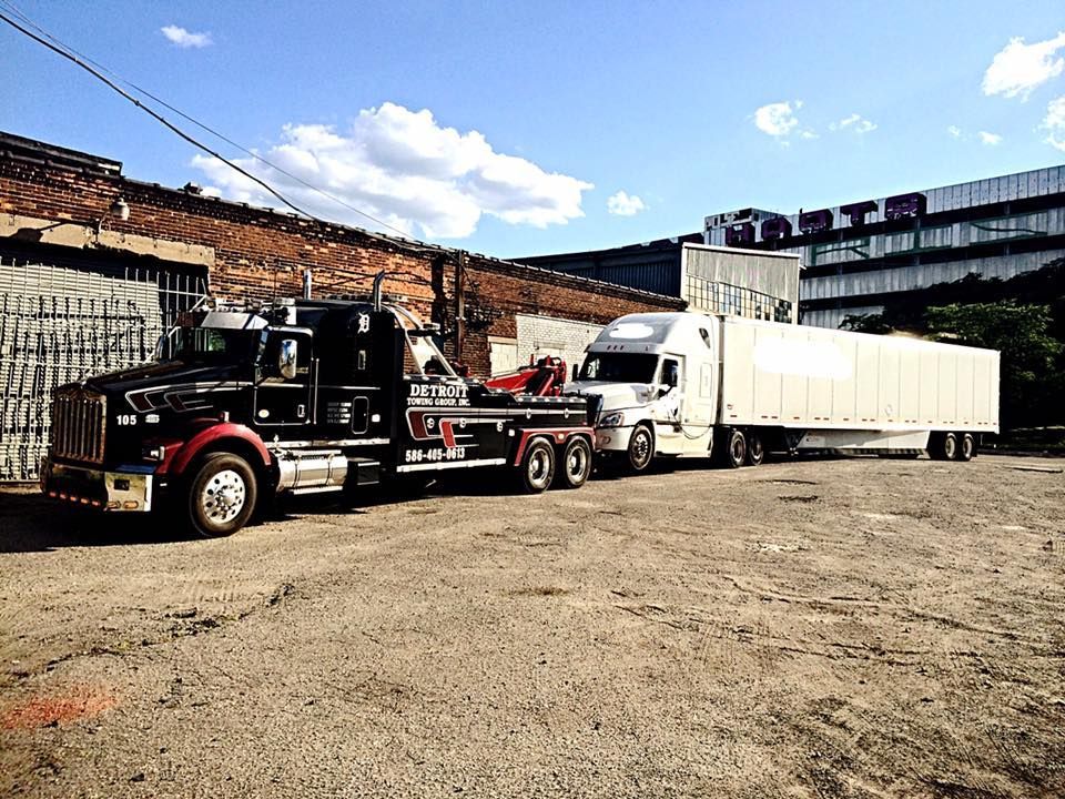 Black tow truck towing a white semi-truck with a long trailer, parked outdoors, near a brick building.