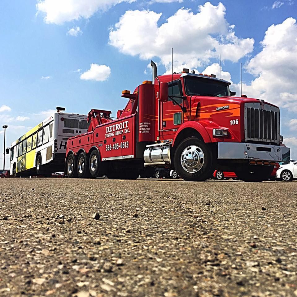 Red tow truck towing a white and yellow bus on a paved lot under a blue sky.