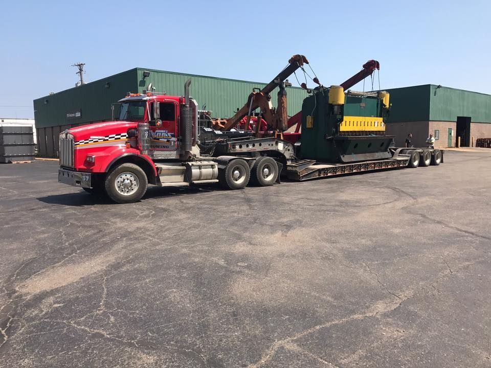 Red semi-truck hauling large green industrial machine on a flatbed trailer in a parking lot.