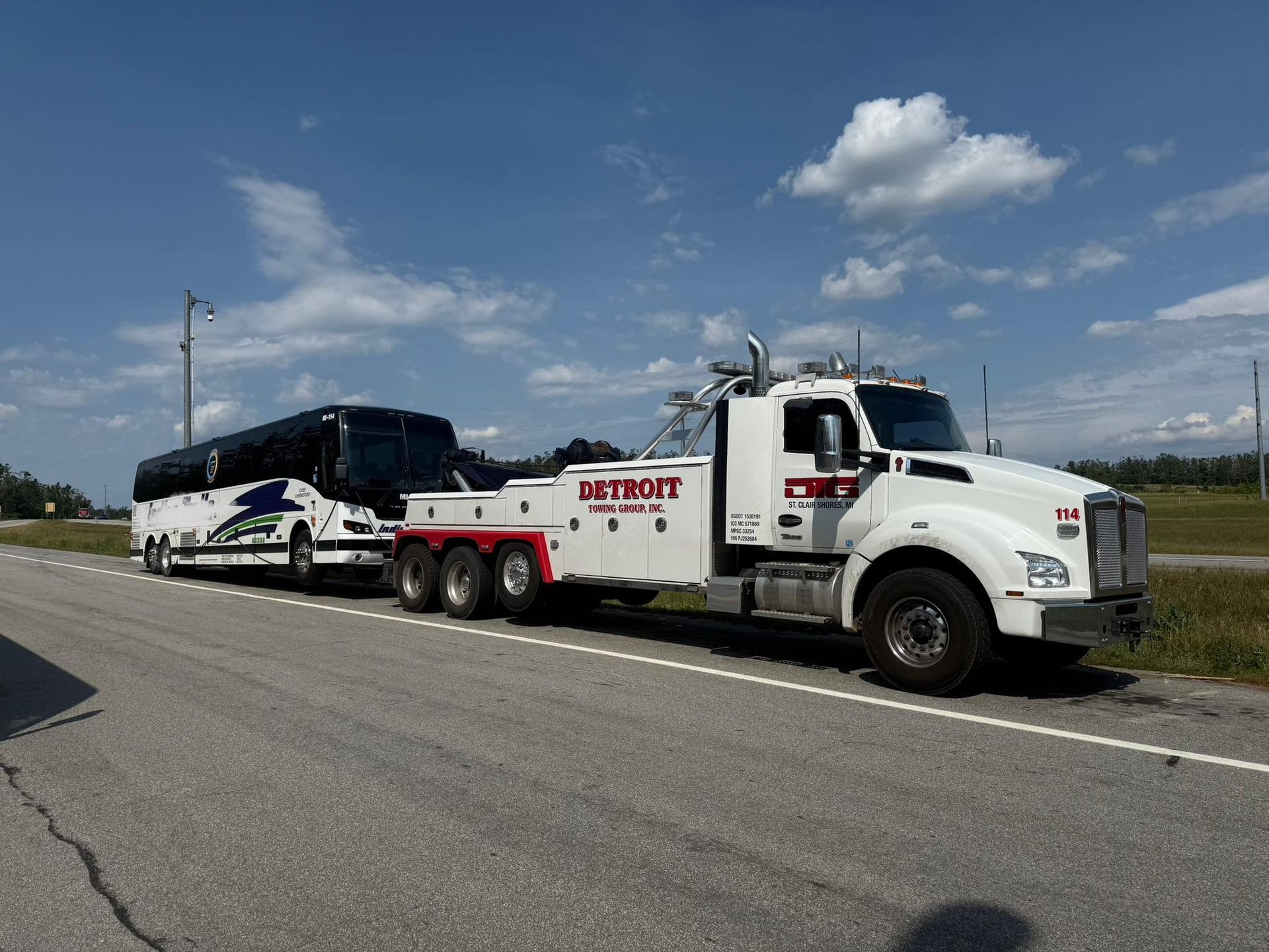 White tow truck towing a white and blue bus on a road with a blue sky.