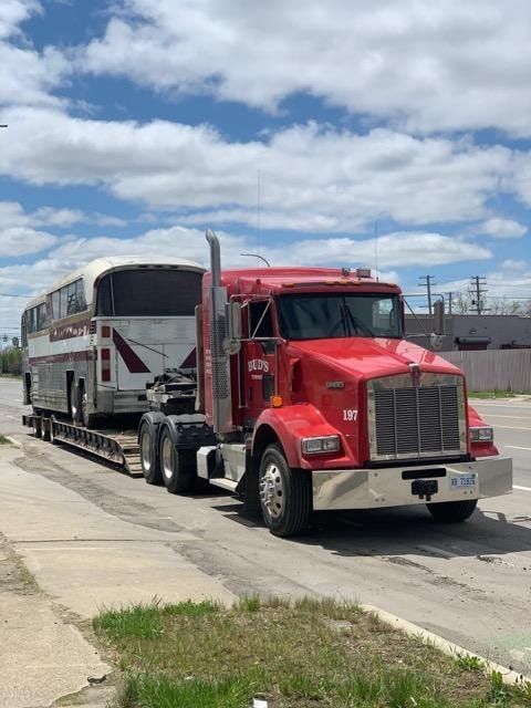 Red Kenworth semi-truck towing a light-colored bus on a flatbed trailer on a paved road, under a cloudy sky.