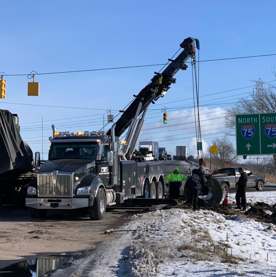 Black tow truck with crane lifting a vehicle from snow-covered roadside near an interstate highway sign.