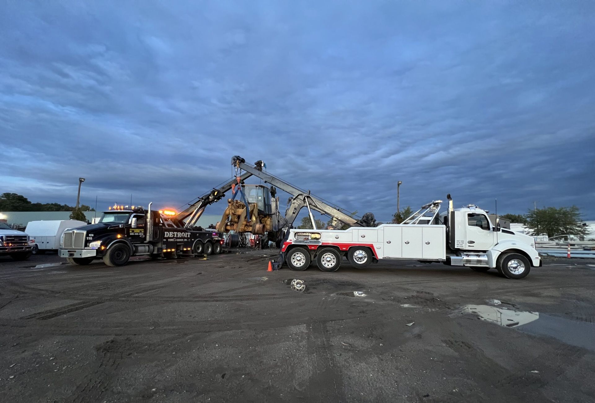 Two tow trucks at a salvage yard, lifting a large construction machine under a cloudy sky.