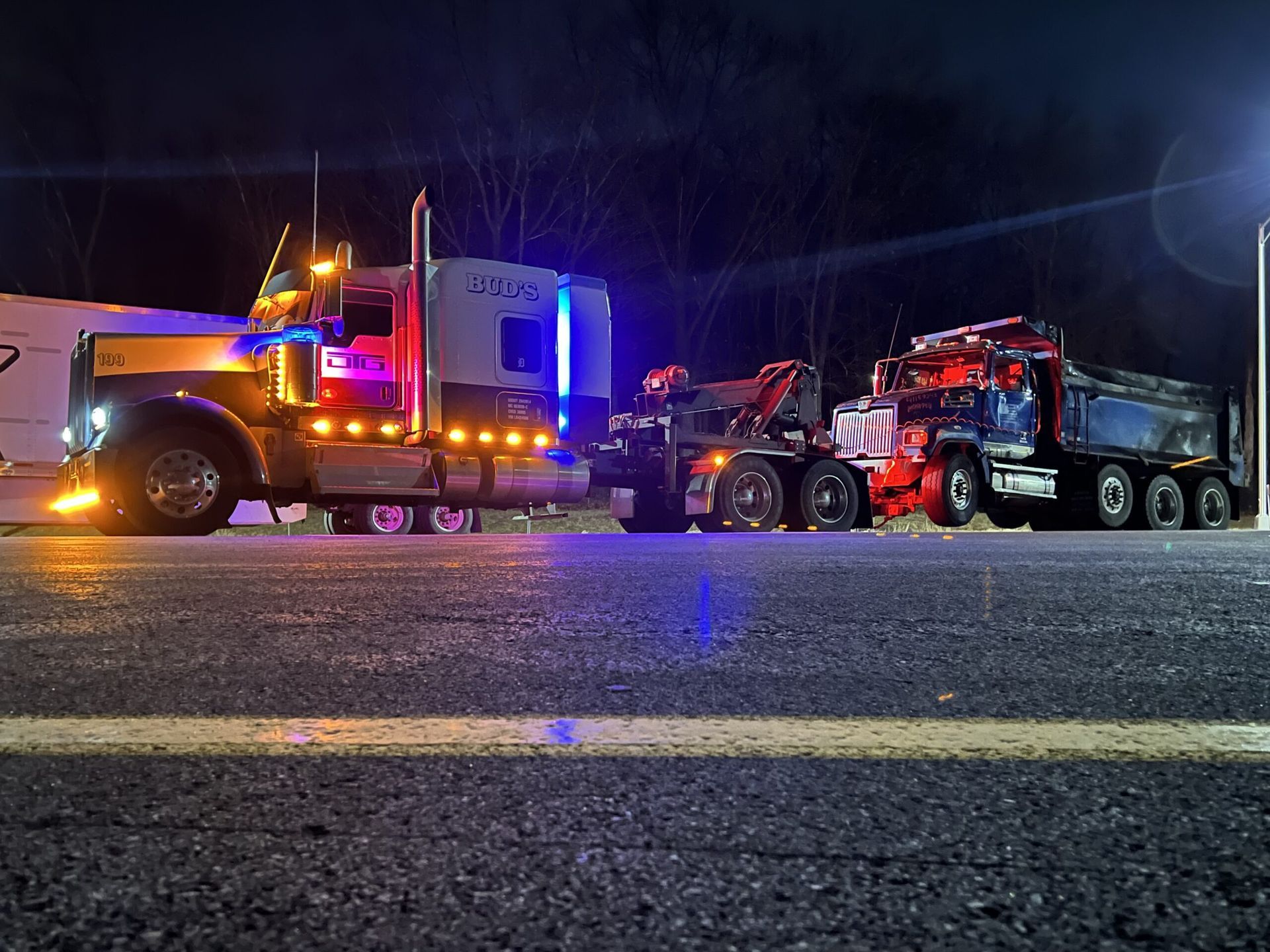 Tow truck pulling a dump truck on a dark road at night; lights flashing, illuminated vehicles.