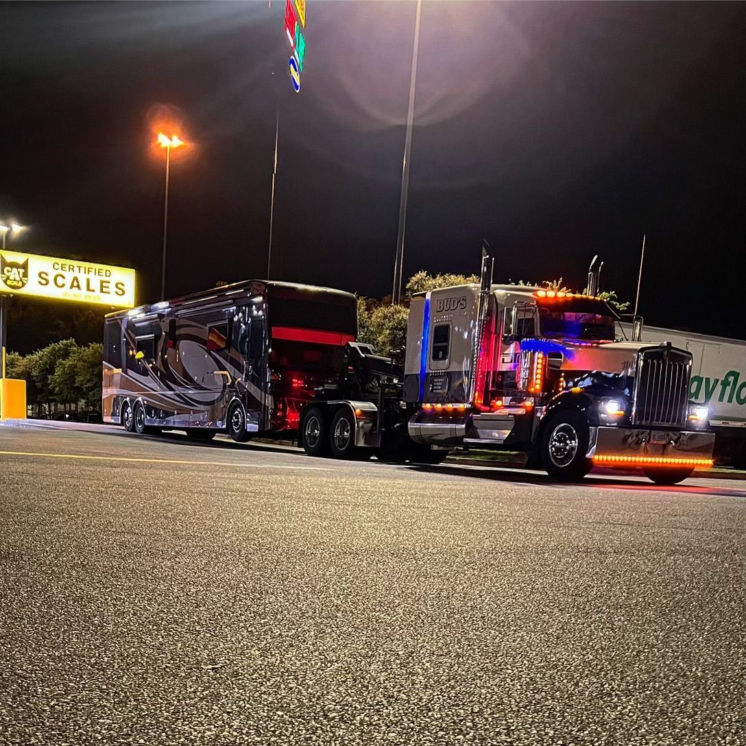 Nighttime shot of a decorated semi-truck hauling a trailer at a truck stop. Bright lights illuminate the vehicles and surrounding area.