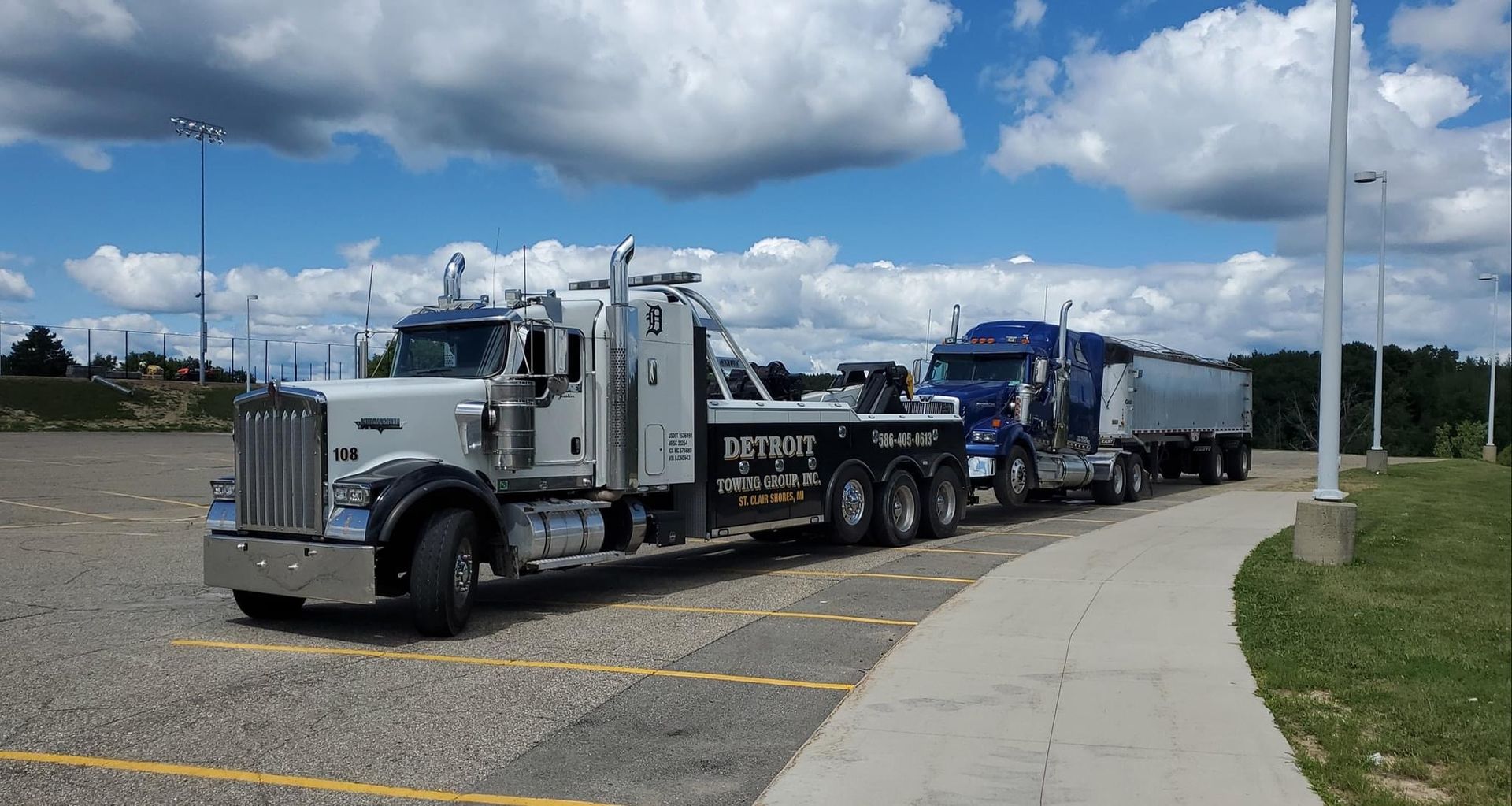 Gray tow truck towing a blue semi-truck with a trailer in a parking lot on a sunny day.