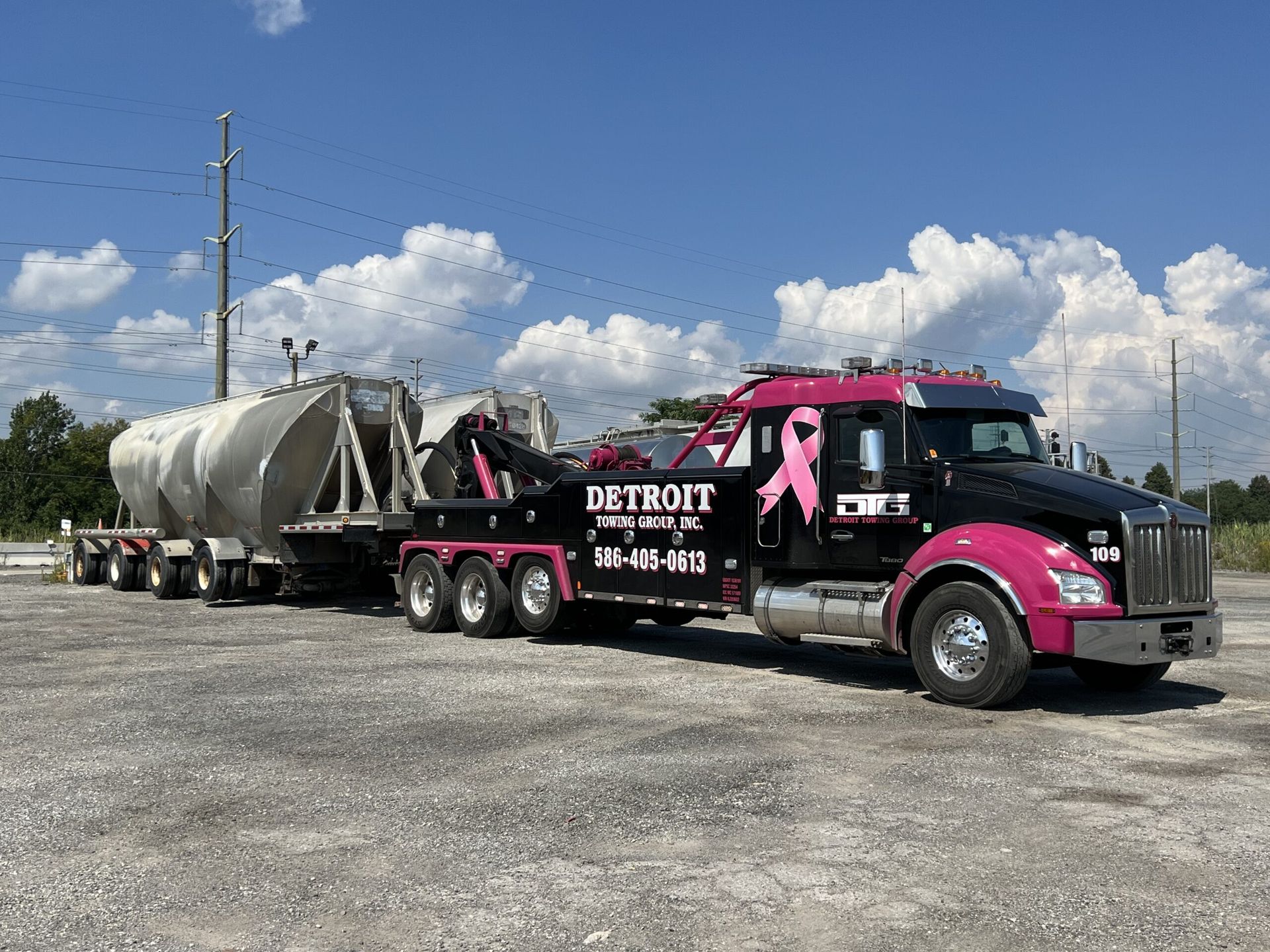 Pink and black Detroit towing truck pulling large silver tank on a flatbed trailer under a blue sky.