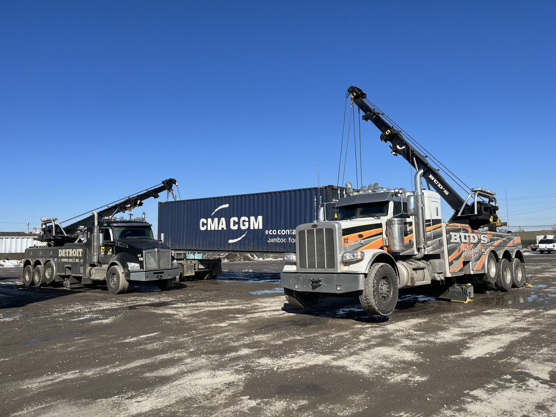 Two tow trucks lifting a blue CMA CGM shipping container on a bright day.