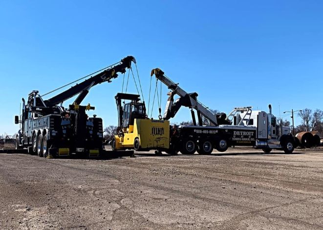 Two tow trucks lifting a yellow forklift on a paved lot under a blue sky.