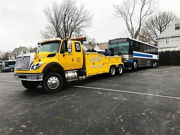 Yellow tow truck towing a blue and silver bus in a parking lot. Cloudy day.