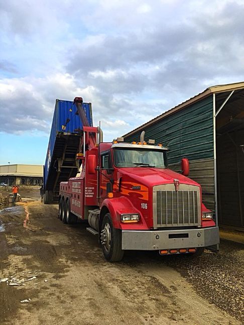 Red semi-truck tilted, lifting a blue shipping container. Outdoors, cloudy sky.