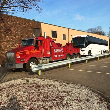 Red tow truck towing a white bus. The truck is parked in a parking lot, near a brick and beige building.