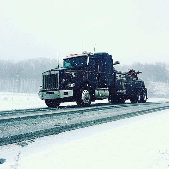 Black tow truck driving on a snow-covered highway during a snowstorm.