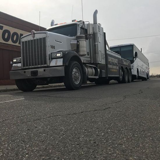 Silver tow truck towing a white bus on an asphalt road in front of a building with a brown facade.