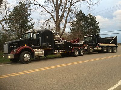 Black tow truck towing a matching dump truck on a road; trees in the background.