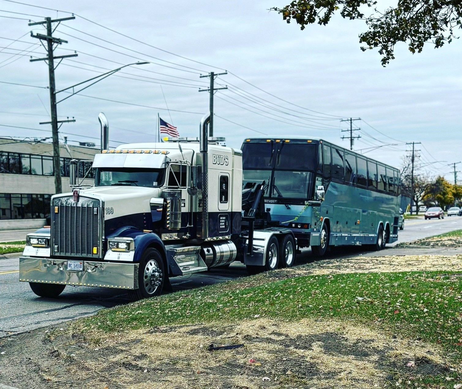 Truck towing a bus on a road with green grass and buildings in the background. Cloudy sky.