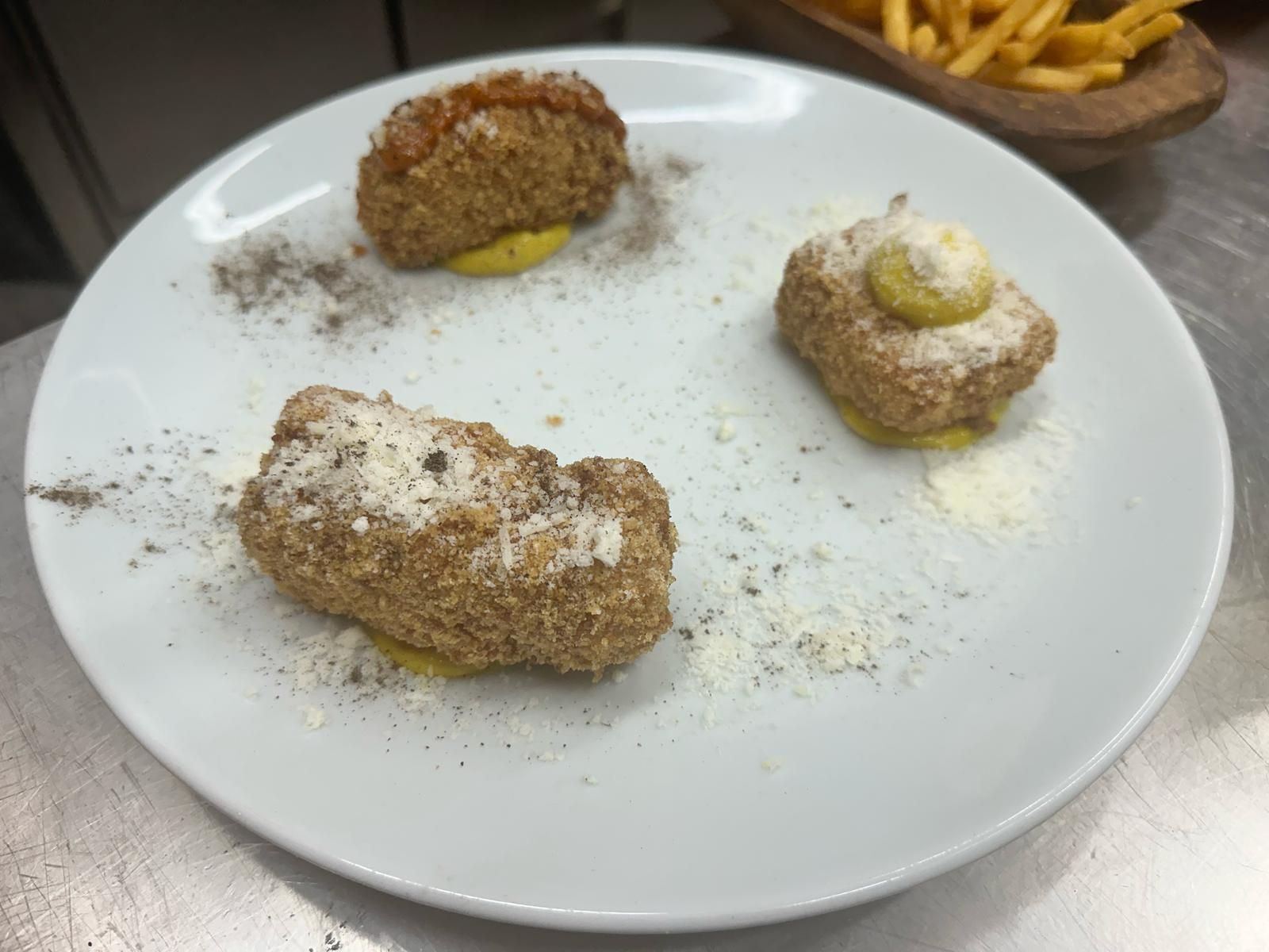 Tre pezzi di cibo impanati e fritti su un piatto bianco, guarniti con formaggio e salsa. Patatine fritte sullo sfondo.