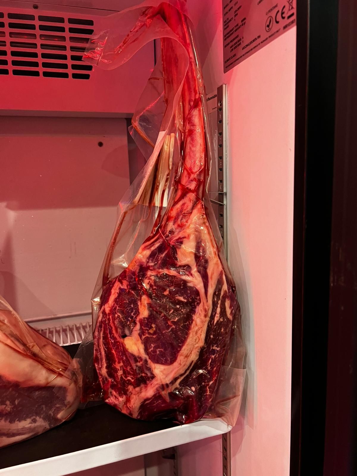 Bistecca tomahawk frollata a secco in un sacchetto sottovuoto, appesa in frigorifero sotto una luce rossa.