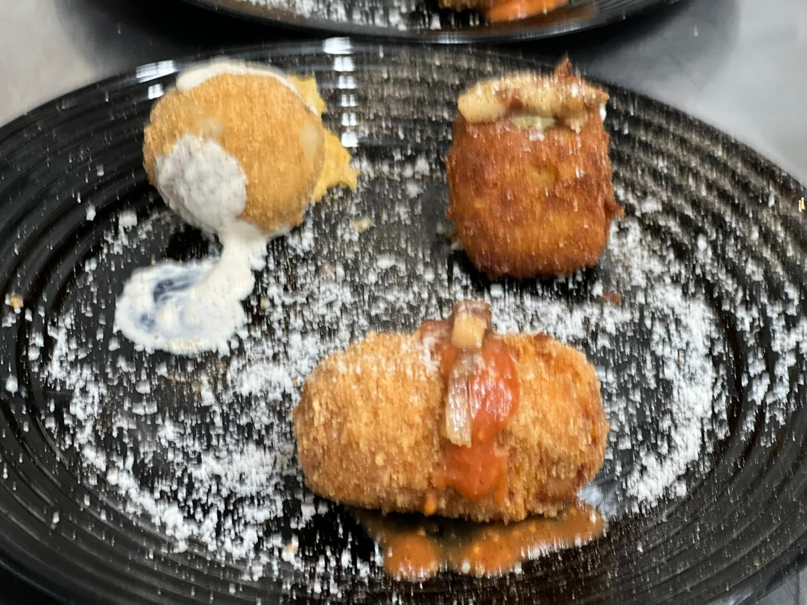 Tre antipasti impanati e fritti su un piatto nero, conditi con salse e spolverati con polvere bianca.