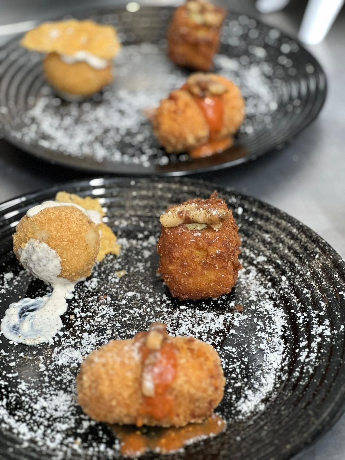 Dolci fritti serviti su piatti neri: rotondi, dorati, ricoperti di salse e zucchero a velo.