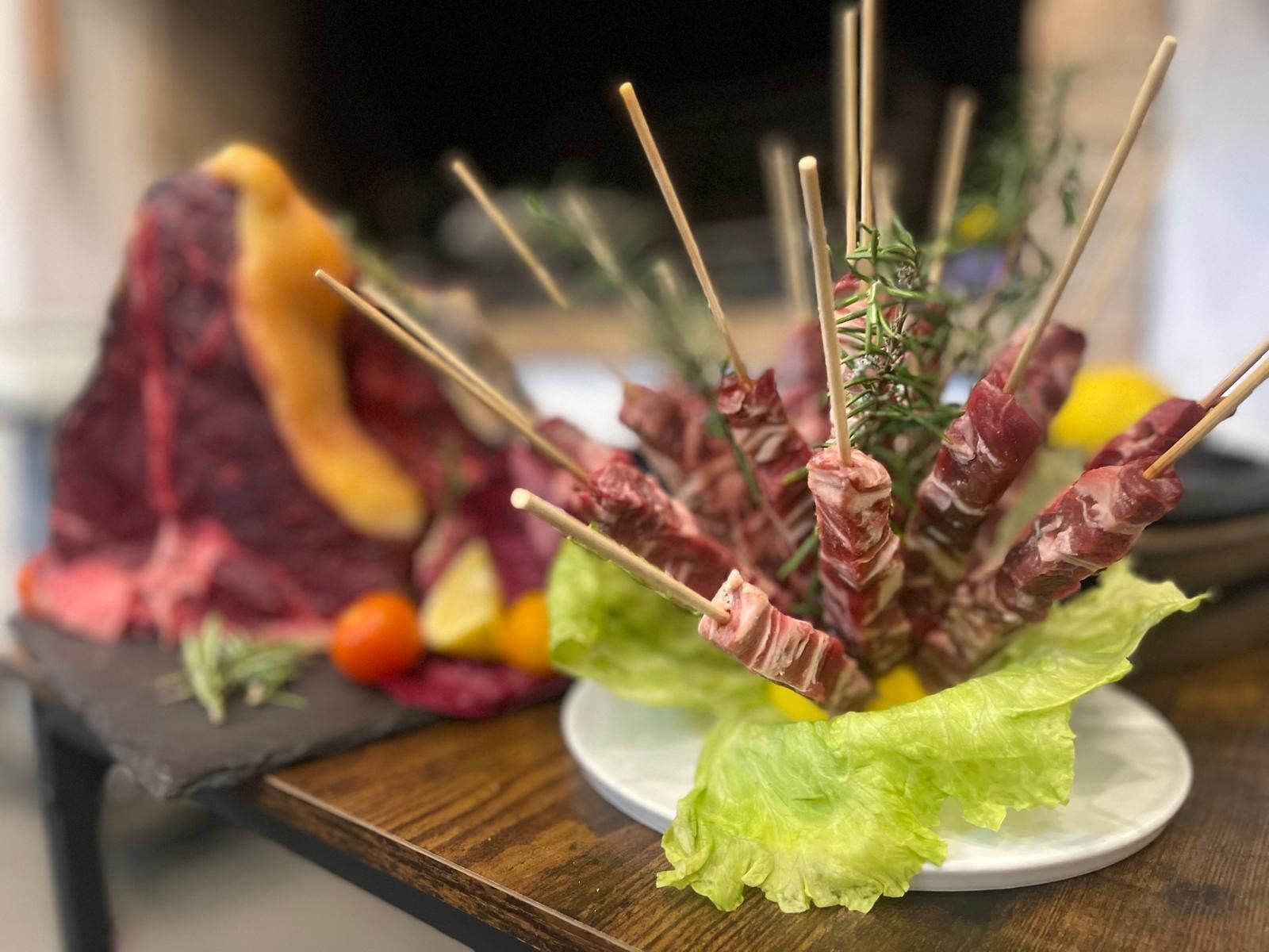 Antipasti di carne infilzata su un piatto bianco con lattuga, rosmarino e una grande bistecca sullo sfondo.