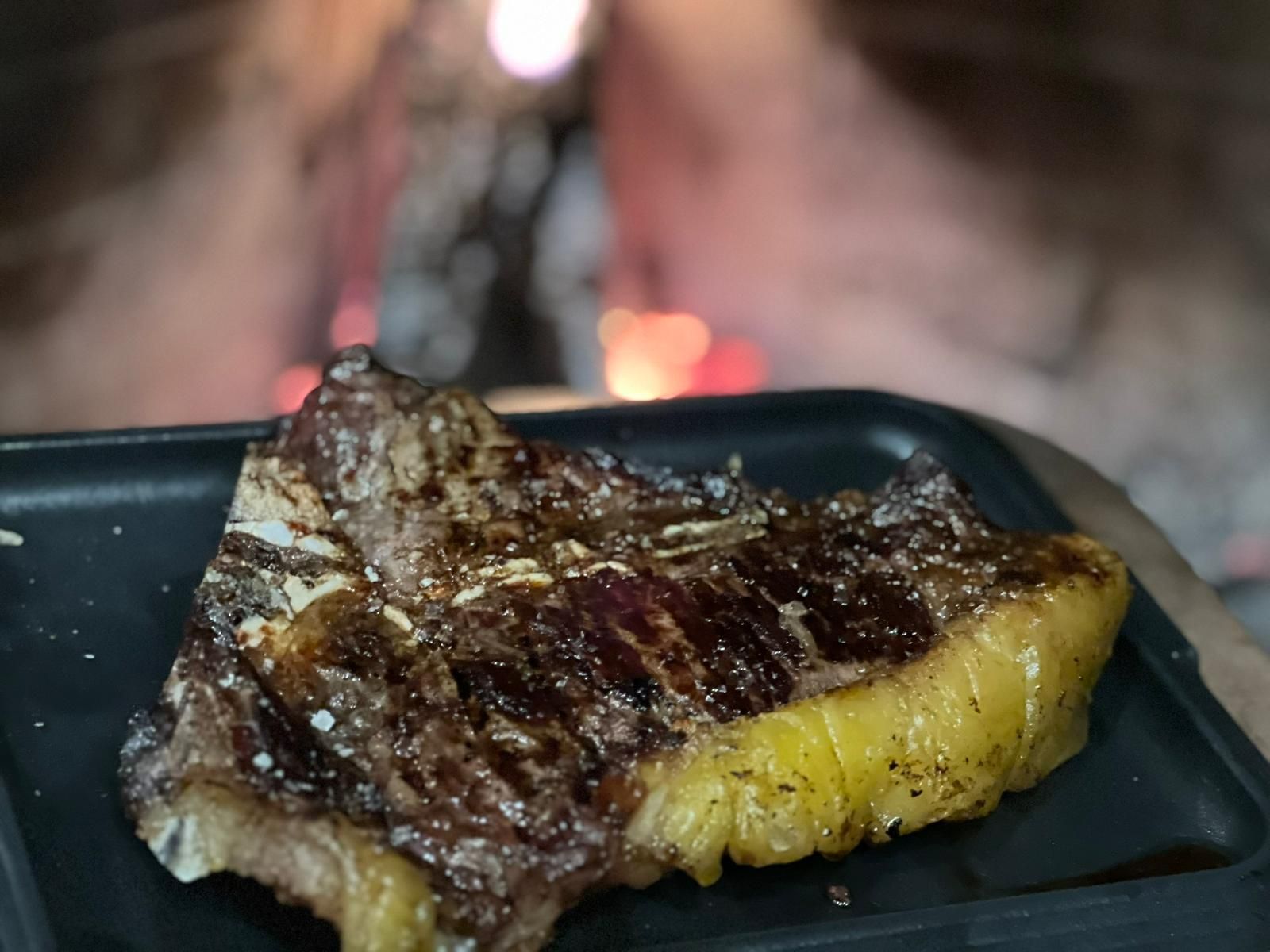 Bistecca alla griglia su un vassoio nero davanti a un forno sfocato con il fuoco.