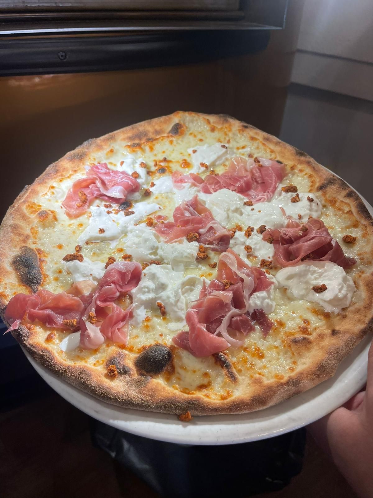 Pizza su un piatto con prosciutto, ricotta e quello che sembra essere pangrattato; sullo sfondo il forno.