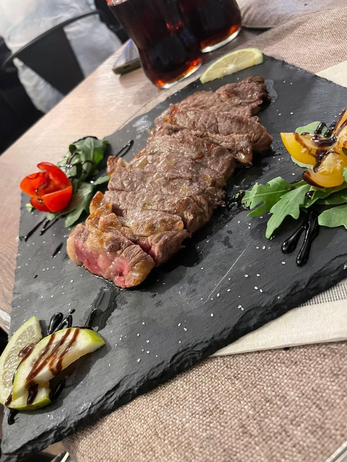 Bistecca servita su un piatto di ardesia nera, con guarnizioni, glassa balsamica e bevande sullo sfondo.