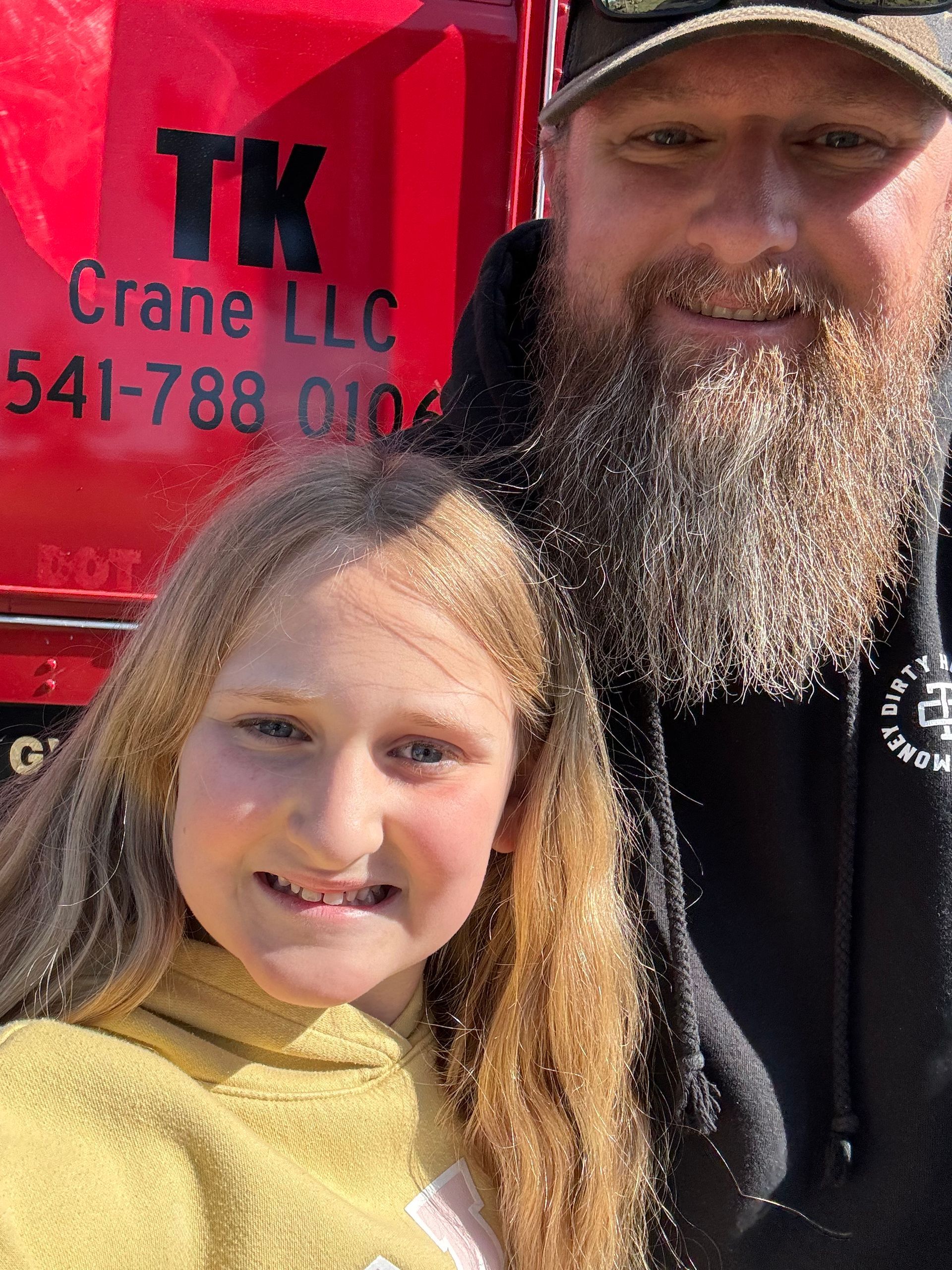 A man with a beard is standing next to a little girl in front of a truck that says tk crane llc