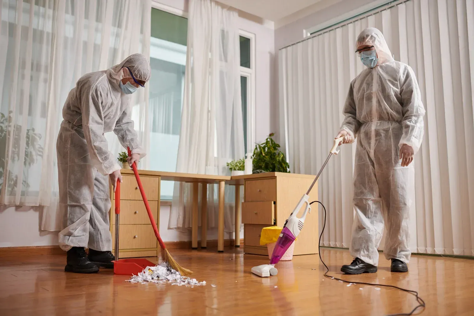 Two people in protective suits cleaning an office, one sweeping debris, the other vacuuming the floor.
