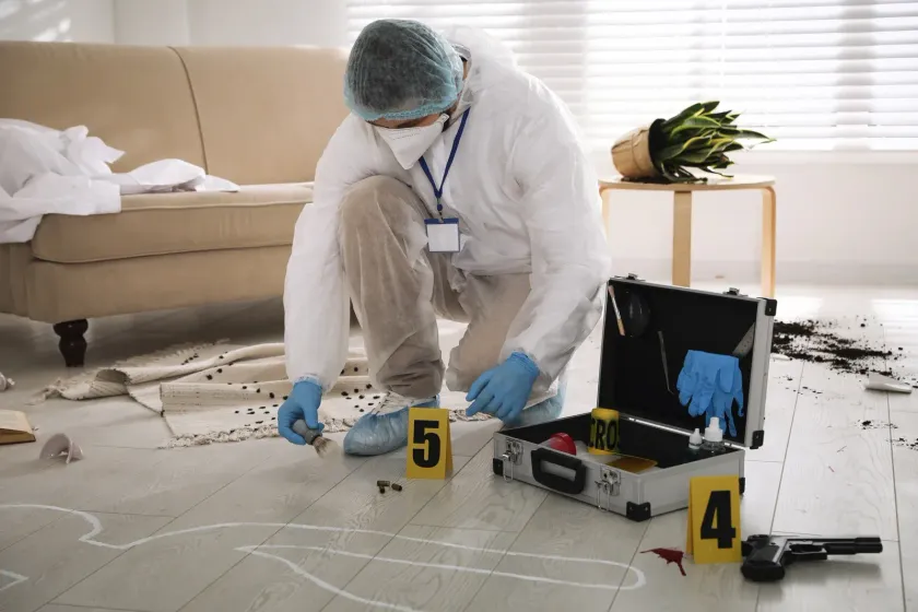 Forensic investigator in protective suit at a crime scene, gathering evidence near a body outline and weapon.
