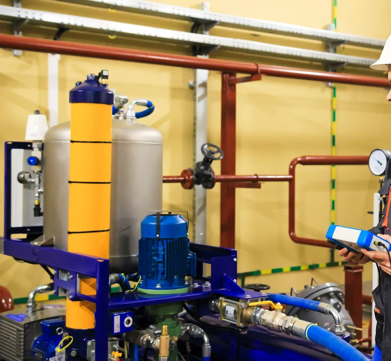 Two workers inspect equipment in an industrial setting. One holds a device; tanks, pipes, and machinery are visible.