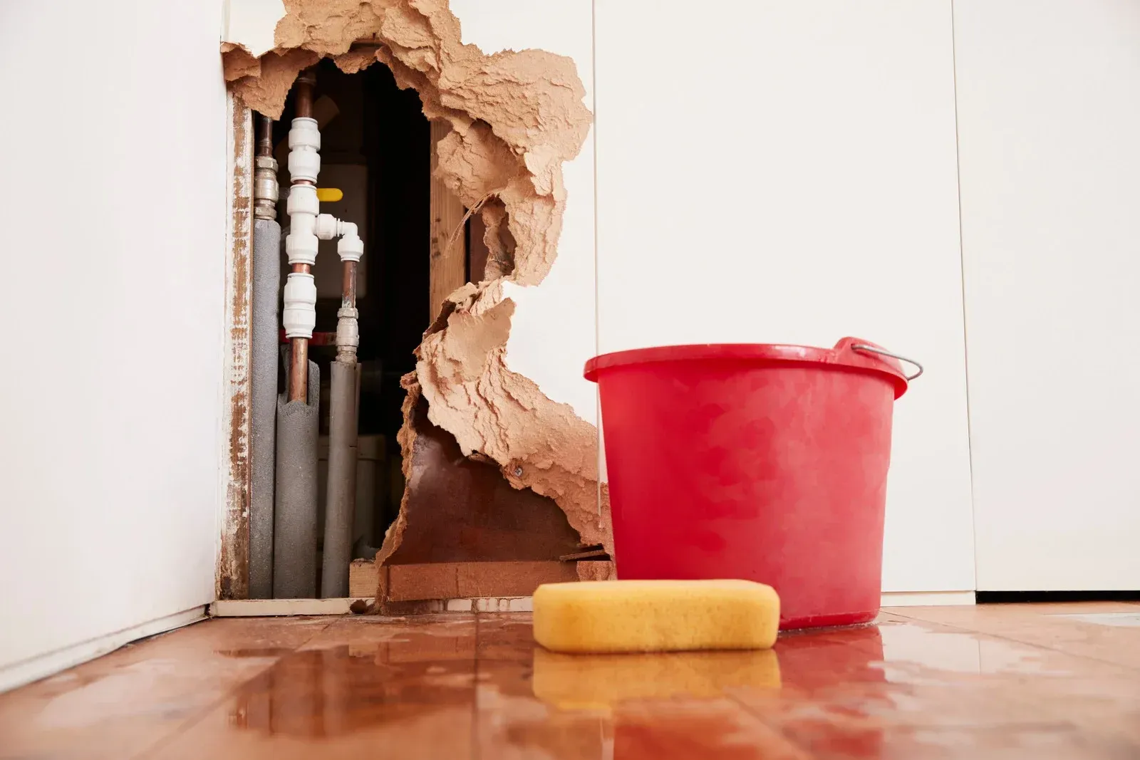Hole in wall revealing pipes, with red bucket and yellow sponge on flooded floor.
