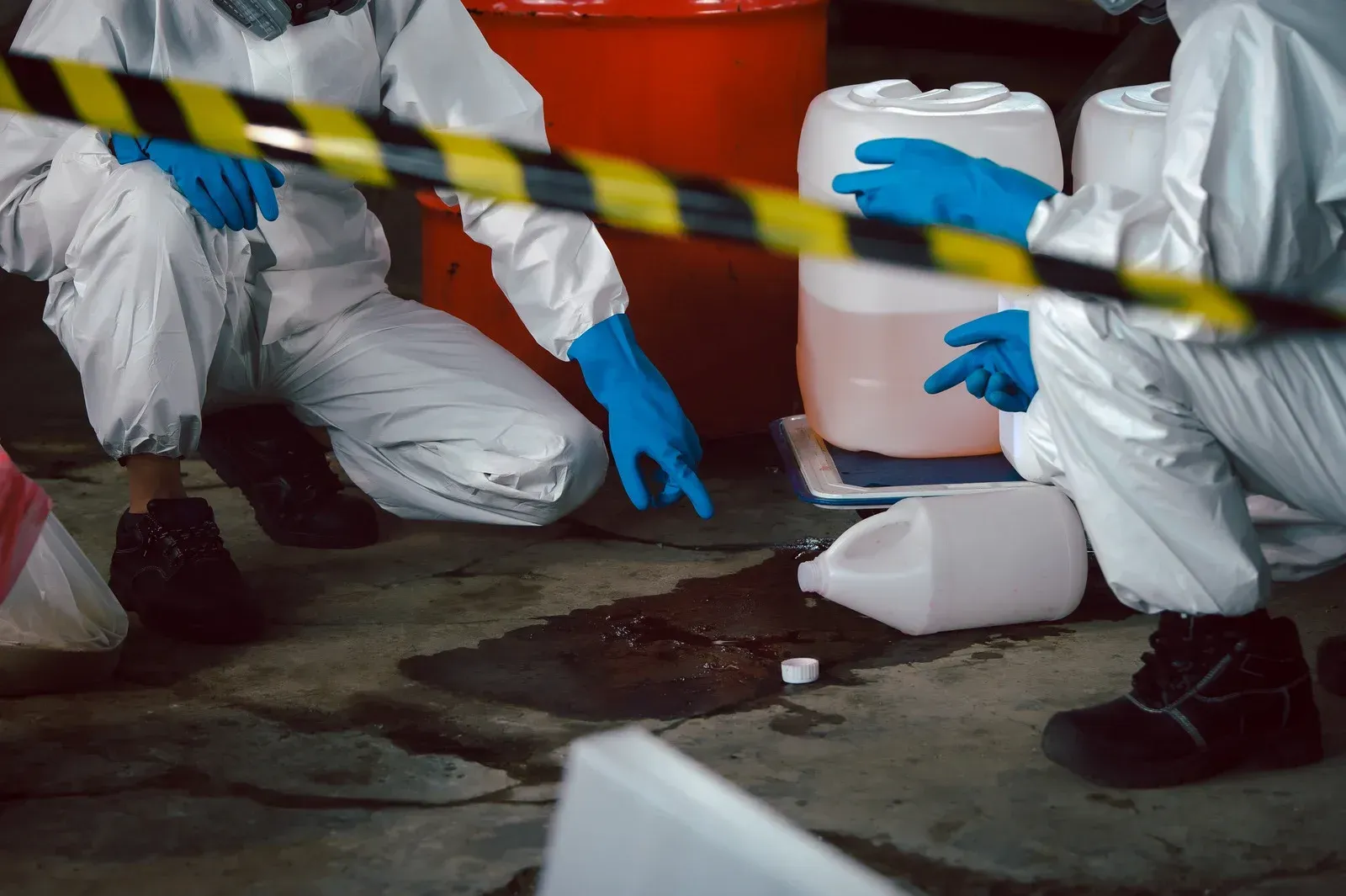 Hazmat-suited individuals inspect a chemical spill on a concrete floor near containers, behind caution tape.