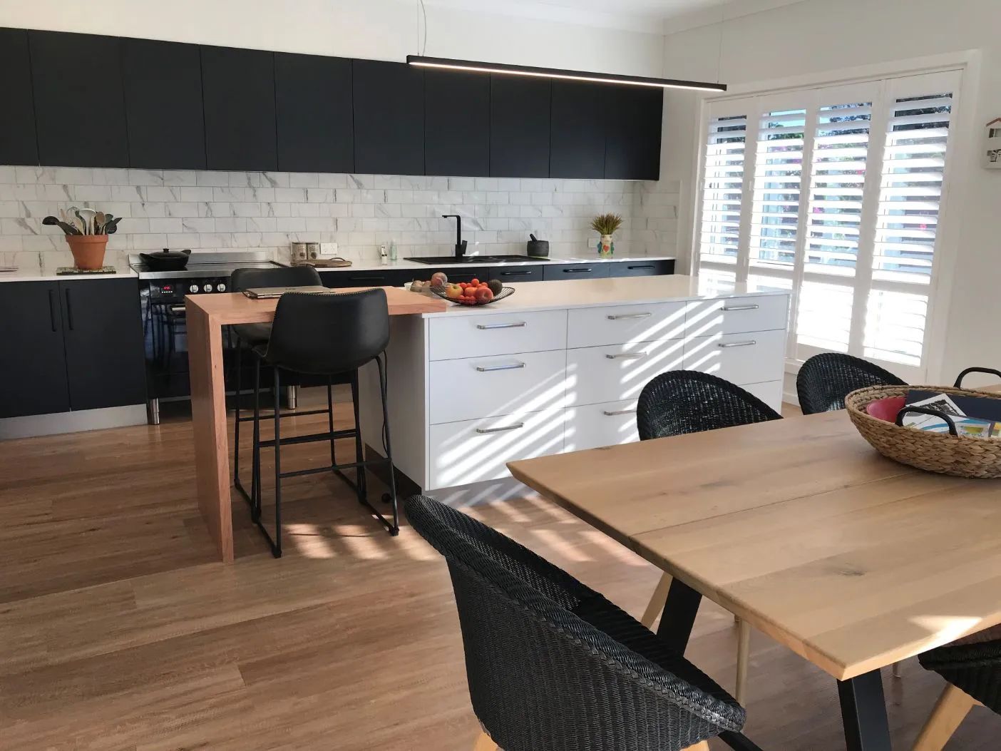 Modern Kitchen With Black Cabinets, White Marble Backsplash, Island With Bar Stools, and a Wooden Dining Table With Chairs — North Coast Tiles in Iluka, NSW