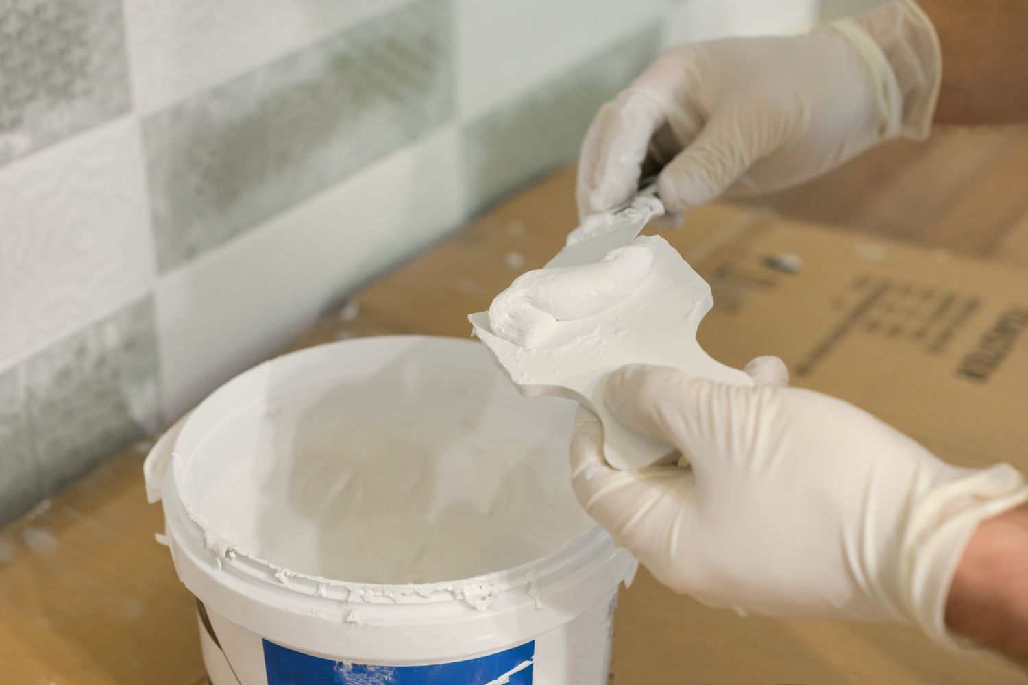 Hands in Gloves Scooping White Material From a Bucket, Likely Grout or Sealant, Against a Tiled Wall Background — North Coast Tiles in Iluka, NSW